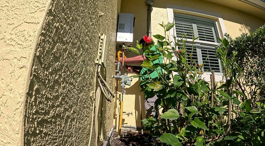 Person inspecting outdoor home plumbing system surrounded by green bushes.