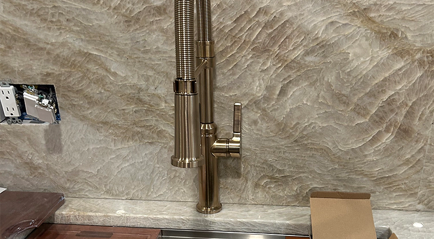 Modern brass kitchen faucet installed with textured stone backsplash.