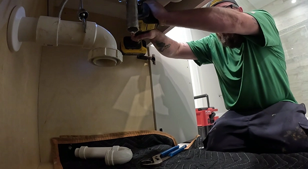 Plumber in green shirt working under sink with tools and pipes visible.