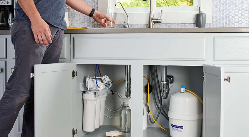 Man inspecting an open cabinet under a kitchen sink with plumbing system.