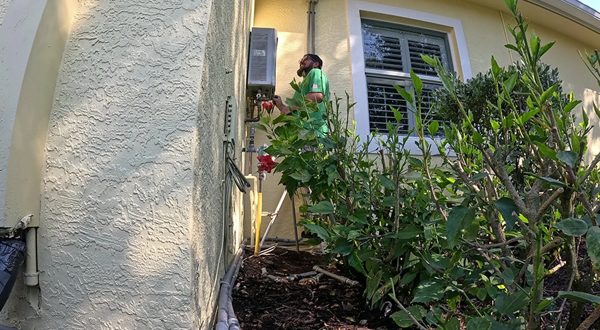 Person inspecting outdoor tankless water heater unit beside a house.