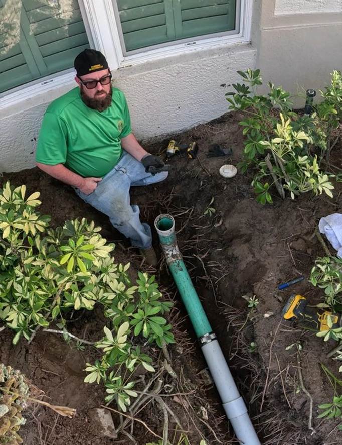 Man in green shirt repairing underground drainage pipe near a house exterior.