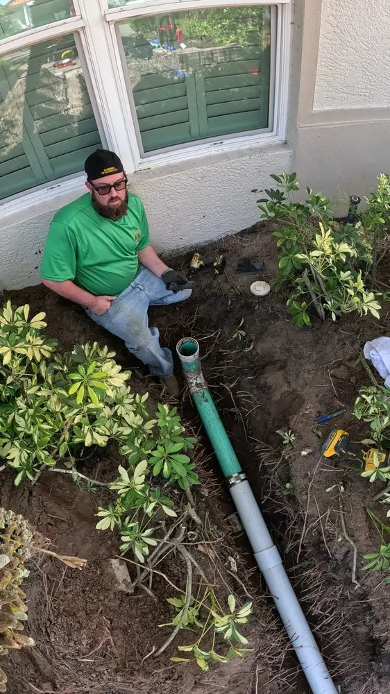 Person in green shirt repairing an outdoor drainage pipe near bushes.