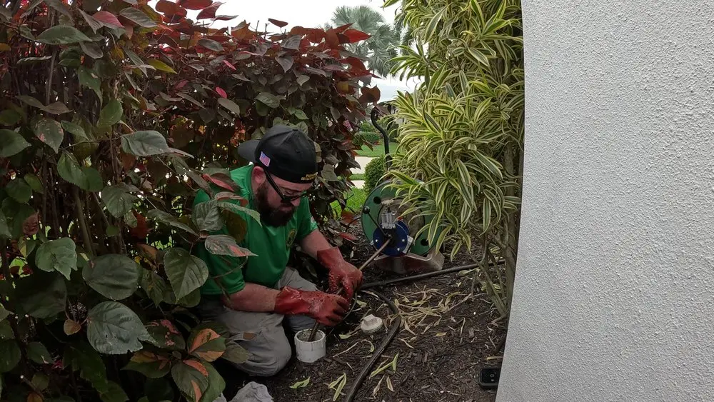 Man in green shirt working on plumbing amidst foliage by a white wall.