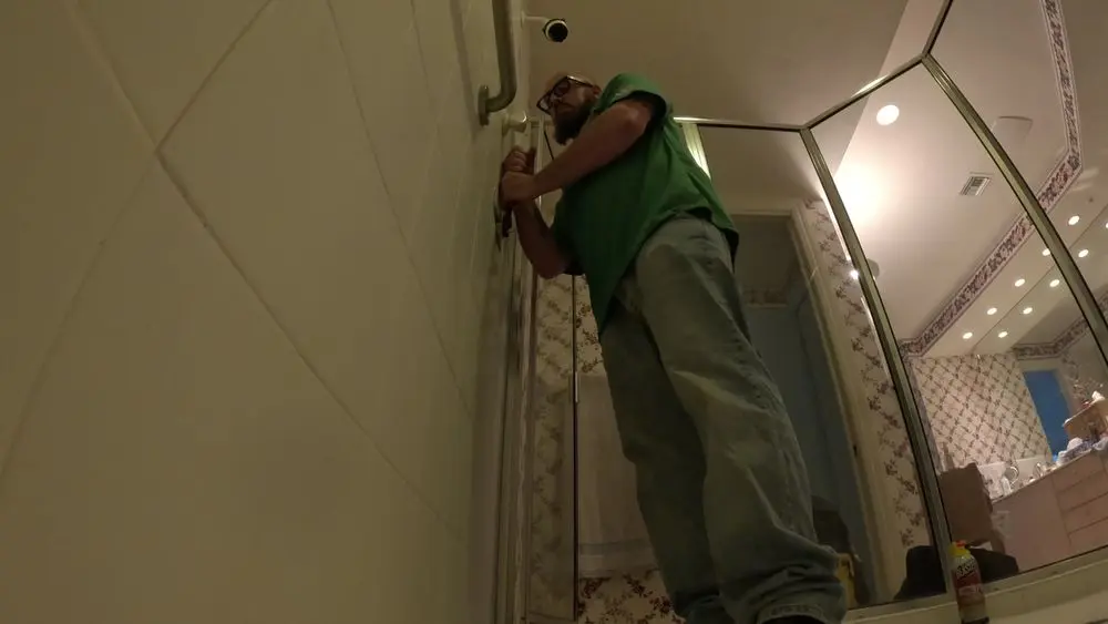 Man in green shirt fixing a showerhead in a bathroom.