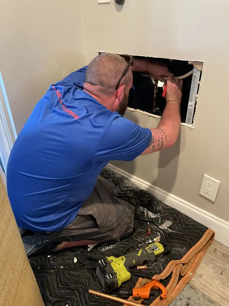 Plumber repairing a pipe inside a wall with tools scattered nearby.