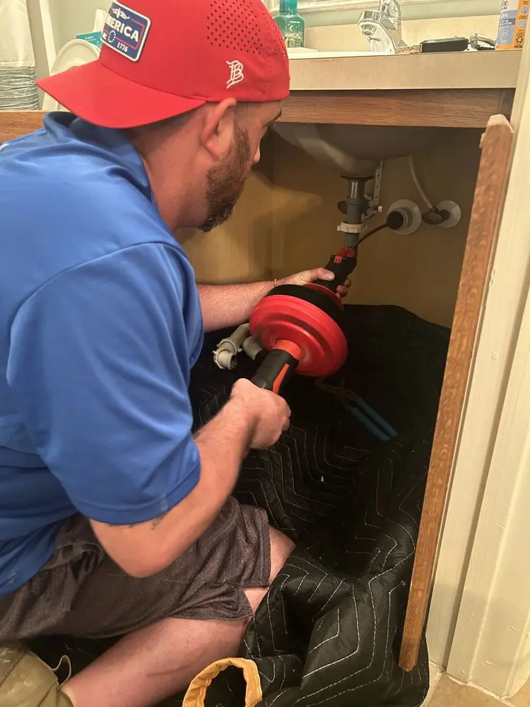 Man in red cap using plumbing tool under sink cabinet.