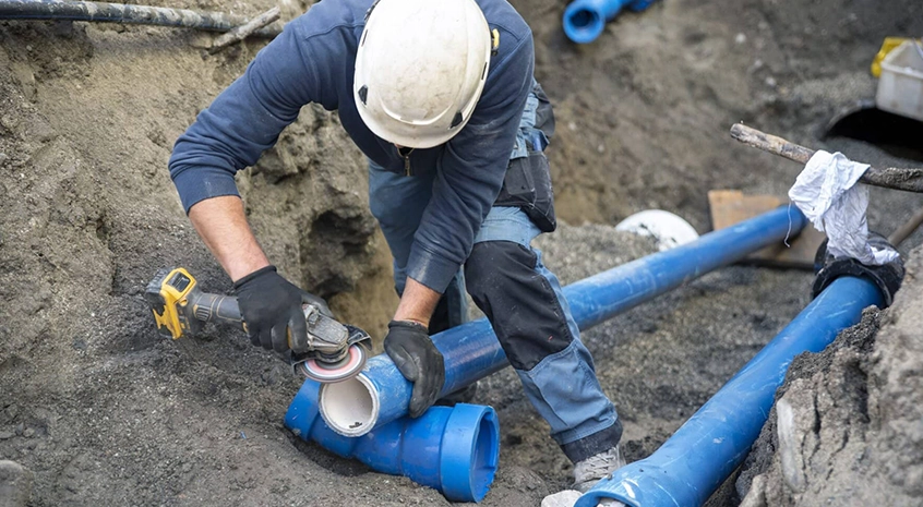 Worker using power tool on blue underground pipes in a trench.