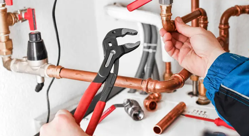 Hands using pliers on copper plumbing pipes with wrench nearby.